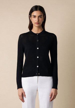 Kobieta o prostych ciemnych włosach, ubrana w czarny, zapinany na guziki kardigan z długim rękawem oraz białe spodnie, stojąca na tle jednolitego beżowego tła.