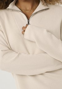 Beżowy sweter z zamkiem błyskawicznym o teksturowanym, prążkowanym wzorze. Cechy to częściowy kołnierz oraz długie, lekko luźne rękawy.