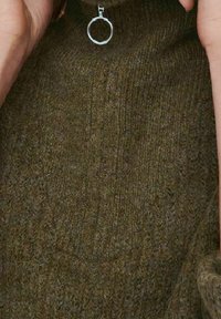 Dzianinowy sweter w oliwkowym kolorze z teksturowanym wykończeniem, z okrągłym srebrnym pierścieniem jako akcentem na dekolcie. Miękki, średniej wagi materiał.
