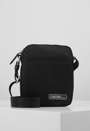 Kompaktowa czarna torebka Calvin Klein na ramię z regulowanym paskiem i przednią plakietką z logo, leżąca na białej powierzchni.