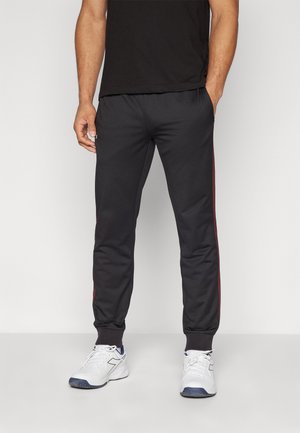 Black athletic pants with red side stripes, elastic waistband, and ribbed cuffs, paired with white sporty sneakers.