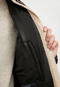 Hånd, der holder en beige jakke åben for at vise dens sorte inderfor med en lynlåslomme og en strikket beige sweater under.