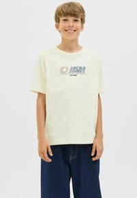Krem majica od pamuka s kratkim rukavima, s grafičkim printom "JACK & JONES" u plavoj i crvenoj boji, u kombinaciji s tamnim traper kratkim hlačama.