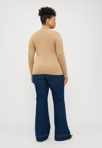 Beige tettsverte genser med høy hals kombinert med blå flagrende jeans. Genseren har lange ermer og en glatt tekstur; jeansene har bølgekant i bunnen.