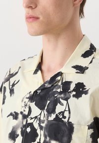 Camicia a maniche corte realizzata in tessuto leggero, con uno sfondo crema e stampa floreale nera, colletto a punta e taschino sul petto.