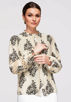 Kvinde justerer manchet på cremefarvet bluse med sorte blomsterprint og blonde detaljer på ærmerne, stående foran en ensfarvet baggrund.