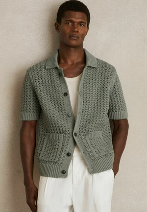 Zelený pletený kardigán s krátkym rukávom a textúrovaným vzorom, s golierovým výstrihom, štyrmi čiernymi gombíkmi a dvomi prednými vreckami na nášivku.