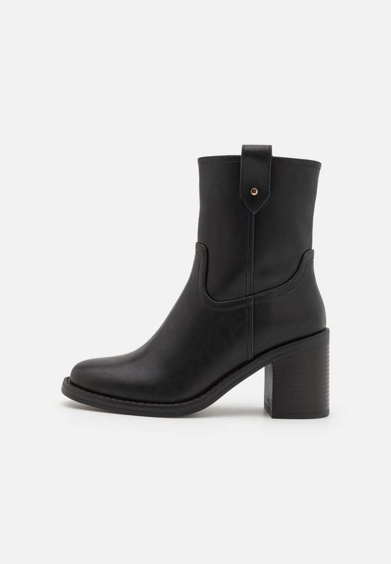 Botte noire en cuir montant jusqu'au milieu du mollet, avec un bout rond, un talon bloc empilé et une languette de tirage ornée d'un rivet doré sur fond blanc.