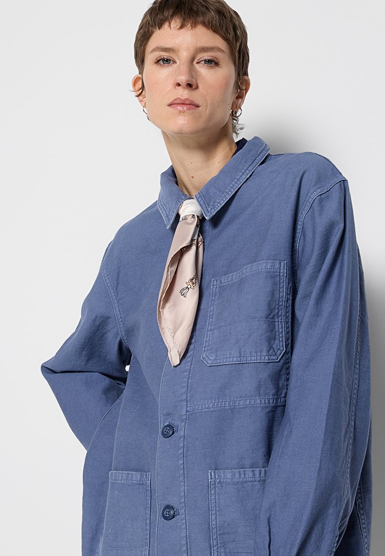 Blaue Baumwolljacke mit Kragen, zwei vorderen Taschen und Knopfverschluss. Ein hellrosafarbenes Tuch ist am Hals gebunden.