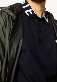 Czarna koszulka polo z kontrastującym białym paskiem na kołnierzyku, zestawiona z ciemnozieloną kurtką zewnętrzną wykonaną z błyszczącego materiału.