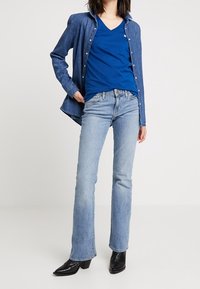 Woman wearing a blue V-neck shirt, unbuttoned denim jacket, light blue bootcut jeans, and black pointed-toe ankle boots standing.