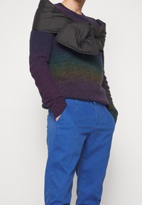 Violet ja sininen gradienttikudossweater, jossa on teksturoitu pinta, yhdistettynä kirkkaan sinisiin housuihin. Musta, pehmustettu yläosa on pukeutunut olkapäiden yli.