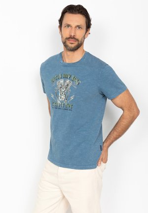 Mand med kort skæg iført en blå T-shirt med grafik og lyse bukser, står med den ene hånd i lommen mod en hvid baggrund.