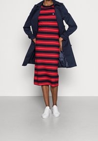 Meriino sininen trench coat päällä punaisia ja meriinossa raidallisia, polveen ulottuvia mekkoja. Valkoiset tennarit. Tasainen kangas vaakaraitojen ja logoyksityiskohtien kanssa.