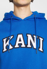 Sudadera azul con capucha, con un gran texto "KANI" bordado en blanco con contorno negro. Hecha de tela suave con cordones ajustables.