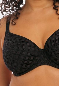 Soutien-gorge noir avec un motif texturé, des bonnets incurvés, des bretelles réglables et une fermeture à crochet et œillet au dos. Tissu doux avec une superposition en maille.