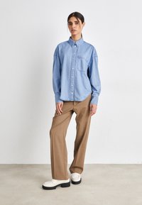 Camicia di denim azzurro chiaro con chiusura a bottoni e tasca sul petto, abbinata a pantaloni ampi marroni e scarpe con plateau bianche. Sfondo neutro.