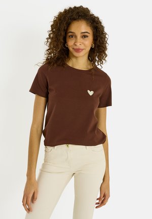 Mujer con cabello rizado, vestida con una camiseta marrón de manga corta con un pequeño emblema de corazón y pantalones beige, posando frente a un fondo blanco liso.