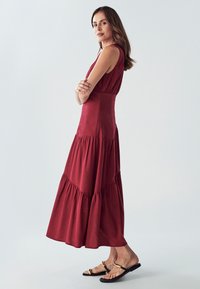 Bordo maxi obleka s večnivojskim dizajnom, izdelana iz mehkega materiala. Ima prilegajoč se zgornji del in pihajočo krilo. Uporabljena z črnimi sandali.