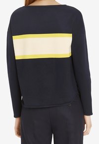 Top lungo navy a maniche lunghe con una larga striscia orizzontale crema e gialla sulla schiena. Tessuto morbido, scollatura rotonda, vestibilità rilassata.