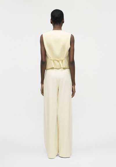 Γυναίκα με σκούρο δέρμα και μαλλιά σε κότσο, φορεί μια μπεζ αμάνικη γιλέκο με πόρπη στο πίσω μέρος και εκρού παντελόνες φαρδιάς γραμμής, στέκεται γυρισμένη μπροστά από την κάμερα.