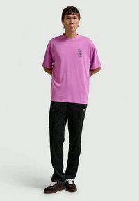 Persoon staat met handen achter de rug, draagt een roze T-shirt met een kleine afbeelding, zwarte broek en donkere sneakers op een effen achtergrond.