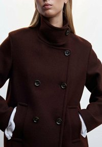 Manteau marron en laine à double boutonnage avec un col montant, doté de boutons noirs et de poches latérales, agrémenté de poignets blancs.