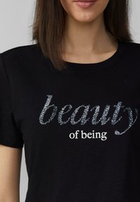 T-shirt noir à col rond avec un texte en sequins argentés disant "beauty" et un texte blanc disant "of being". Matériau en coton doux.