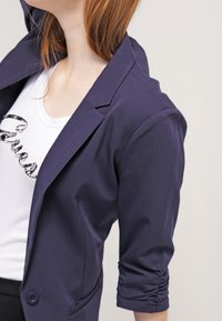 Granatowa marynarka o dopasowanym kroju, z rękawami o długości trzech czwartych i marszczonym detalem, noszona na białej koszuli z czarnym nadrukiem graficznym.