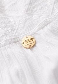 Tessuto bianco con un motivo testurizzato e pieghe, caratterizzato da un'etichetta circolare dorata con la scritta "Superdry BRAND."
