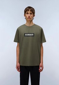 Bawełniany t-shirt w oliwkowo-zielonym kolorze, z okrągłym dekoltem, krótkimi rękawami i czarnym, prostokątnym logo umieszczonym na środku klatki piersiowej.