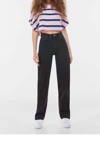 Svarta högmidjade jeans med rak benpassform och fyra fickor, kombinerat med en kort topp med randig rosa och marinblå design. Vita sneakers är på. 