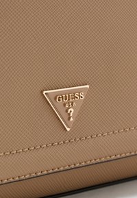 Primo piano di una borsa beige testurizzata, caratterizzata da un logo dorato triangolare "GUESS" con un punto interrogativo, accentuato da cuciture dettagliate.