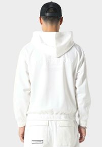 Felpa bianca con cappuccio, dal taglio rilassato, con polsini e vita elasticizzati. Presenta un logo discreto sulla schiena. Indossata con un berretto nero.