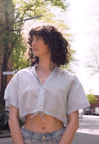 Camisa azul clara, de botões, cropped, com riscas brancas verticais e mangas curtas. Amarrada na bainha; combinada com calções de ganga de cintura alta.