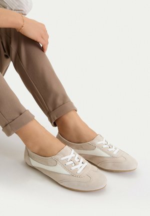 Person trägt beige-weiße Schnürschuhe im Freizeitstil mit hochgekrempelten braunen Hosen vor einfarbigem Hintergrund.