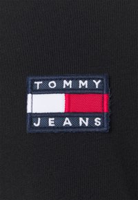 Svart tygmärke med texten "TOMMY" i vitt och "JEANS" i vitt på en blå bakgrund, med ett horisontellt rött och vitt rand.