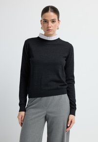 Zwarte trui met een geribde halslijn en een witte, gerimpelde kraag. Lange mouwen en een aansluitend lijfje, gecombineerd met grijze broek met spijkerstreep.
