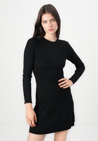 Musta ribattu pitkähihaine mekko, jossa on tiukka siluetti ja pyöreä takaosa. Ominaisuuksiin kuuluu pystysuorat raidat ja lievä levennys helmassa.