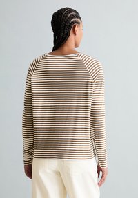 Frau mit geflochtenem Haar, die ein braun-weiß gestreiftes Langarmshirt und cremefarbene Hosen trägt, von hinten vor hellem Hintergrund.