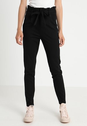 Femme portant un pantalon noir taille haute avec une ceinture nouée, associé à des baskets rose clair et un top blanc sans manches.