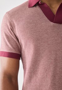 Camisa polo de manga corta con un patrón texturizado, rosa con un cuello y puños en burdeos oscuro. Diseño de cuello en V, acabado de tela suave.