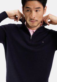 Pull confortable à demi-zip de couleur marine en tissu tricoté doux, avec un logo subtil sur la poitrine et porté par-dessus une chemise col rose clair.