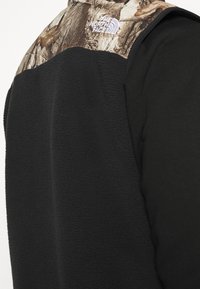 Czarny polarowy kamizelka z kamuflażowym wzorem w okolicy szyi i ramion, z białym logo po lewej stronie. Teksturowany materiał zwiększa ciepło.