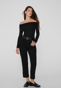 Schwarzes Oberteil mit schulterfreier Optik und Raffung, kombiniert mit hoch taillierten schwarzen Hosen und schwarzen Slippern. Einfaches und figurbetontes Design.
