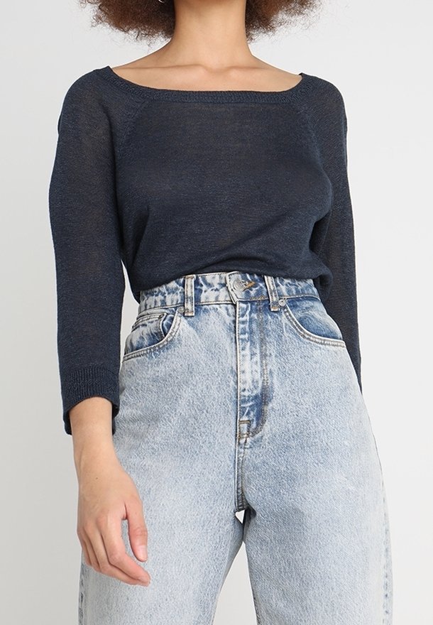 Femme portant un haut bleu marine à manches longues et épaules dénudées, rentré dans un jean en denim clair taille haute, sur un fond uni.