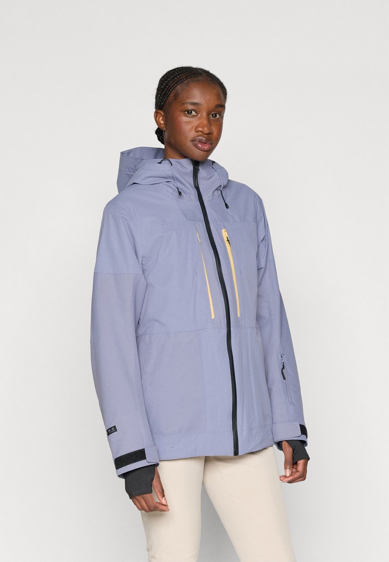 Valkoinen vedenpitävä takki, jossa on huppu, mustat vetoketjut ja keltaiset vetoketjulliset taskut. Varustettu säädettävillä hihoilla ja sileällä pinnalla.