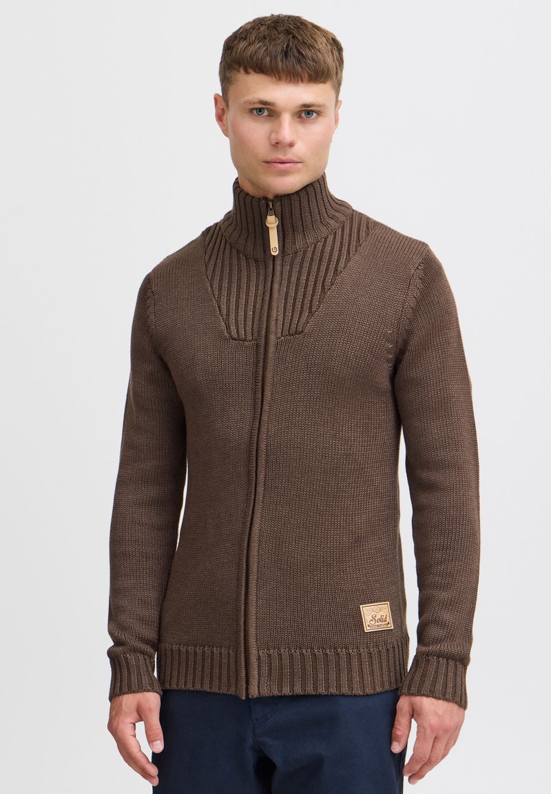 Maglione lavorato a maglia marrone con colletto alto, dotato di chiusura a zip e dettagli a coste sul collo e sui polsini. Toppa con logo sul lato.