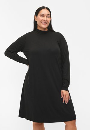 Mujer sonriendo con cabello largo y oscuro, vestida con un vestido negro de manga larga y hasta la rodilla, con una mano en la cadera, contra un fondo blanco liso.