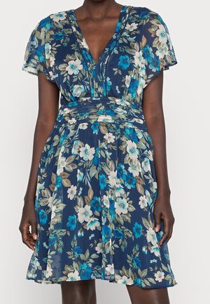 Mørkhudet person iført en marineblå knælængde kjole med hvidt, turkis og lysebrunt blomstermønster og korte flagrende ærmer.